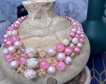 May include: A multi-strand necklace with pink and white beads, displayed on a wooden form. The necklace features various sizes of pink and white beads, interspersed with clear crystal-like beads. The wooden form is light brown, and the background is a marbled gray.