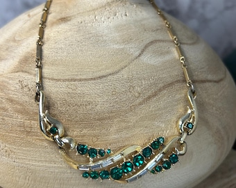 May include: A vintage gold-tone necklace with emerald green rhinestones. The necklace features a delicate chain and a central design with a curved shape, adorned with round and baguette-cut green stones. The necklace is displayed on a wooden vase.