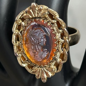 May include: A vintage gold-toned ring featuring an oval amber-colored cameo with a profile of a woman. The cameo is set within a decorative gold filigree frame. The ring is a classic piece of jewelry.
