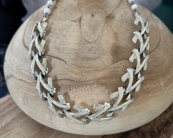 May include: A silver chain necklace with a geometric design. The chain is made of interlocking links that form a pattern of triangles. The necklace is displayed on a wooden surface.