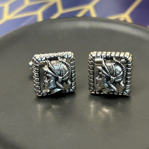 May include: A pair of square silver cufflinks. Each cufflink features a detailed profile of a warrior in a helmet within a decorative frame. The cufflinks are displayed on a dark gray surface.