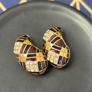 May include: A pair of vintage gold-toned earrings. Each earring features a geometric pattern of black, burgundy, and gold enamel, accented with clear rhinestones. The earrings have a teardrop shape and are set on a dark grey surface.