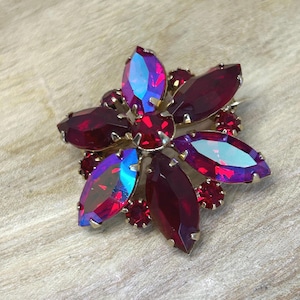 Puede incluir: Broche en forma de flor con engaste dorado. El broche presenta grandes cristales rojos oscuros tallados en marquesa y cristales rojos redondos más pequeños. Algunos cristales tienen un brillo iridiscente azul-violeta. Joya de estilo vintage.