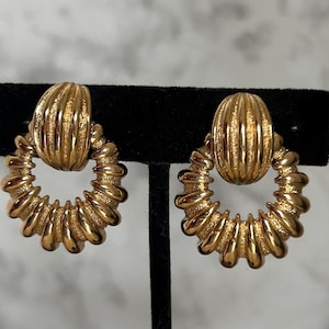 May include: A pair of gold-coloured earrings. Each earring has a textured, oval-shaped top and a circular, ridged design. The earrings are displayed on a black stand against a blurred background.