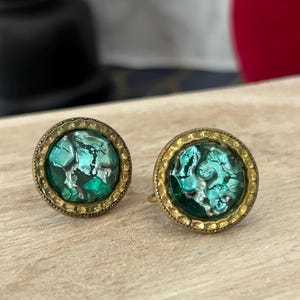 May include: A pair of vintage stud earrings. Each earring features a round, iridescent teal and green center stone set in a gold-toned, textured frame. The earrings are displayed on a light wood surface.