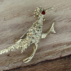 May include: A gold-toned brooch in the shape of a roadrunner with a red rhinestone eye. The brooch is covered in small rhinestones.
