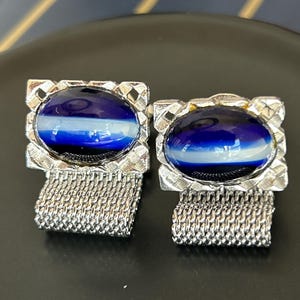 Puede incluir: Un par de gemelos vintage con un engaste de metal plateado. Cada gemelo presenta una piedra ovalada de vidrio azul y blanco en capas. Los gemelos tienen un accesorio de cadena de malla.