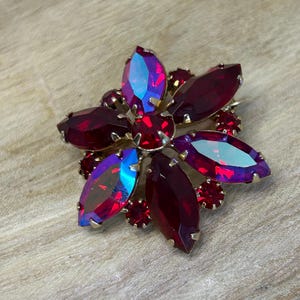 Op de afbeelding: Een bloemvormige broche met een goudkleurige zetting. De broche heeft grote, dieprode, ovaal geslepen edelstenen met iriserende blauwe en paarse highlights. Kleinere rode edelstenen zijn gegroepeerd rond het midden en tussen de grotere stenen.