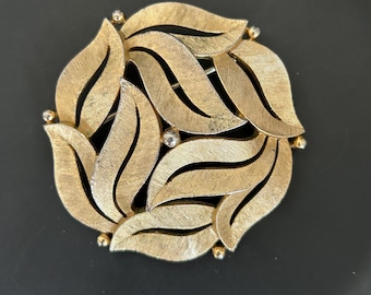May include: A gold-toned vintage brooch with a leaf design. The brooch features overlapping textured leaves arranged in a circular pattern. Small gold-colored beads are placed around the edges. A classic piece of jewelry.