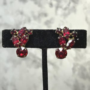 Peut inclure: Une paire de boucles d'oreilles à clip composées de grappes de strass facettés rouge foncé montées sur du métal doré. Chaque boucle présente une combinaison de pierres taillées en marquise et rondes disposées en motif floral avec une pierre ronde à la base.