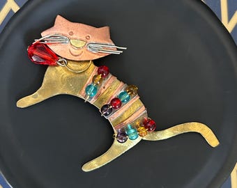 May include: A whimsical cat-shaped brooch made of brass, copper, and colorful beads. The cat has a copper head with silver whiskers and a red crystal in its mouth. The body is adorned with a beaded collar of various colors.