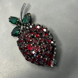 May include: A vintage brooch in the shape of a strawberry. The brooch is covered in red rhinestones, with green rhinestone leaves and a silver-tone stem. The jewelry piece is set against a gray background.