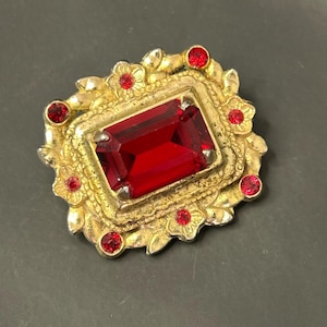 Könnte beinhalten: Eine goldfarbene Brosche mit einem großen roten Edelstein in der Mitte, umgeben von kleineren roten Edelsteinen und einem floralen Design.