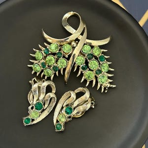 May include: A vintage jewellery set comprising a large brooch and a pair of earrings. The brooch is shaped like a stylised leaf with a swirl design and is adorned with green rhinestones. The earrings are also leaf-shaped and feature green rhinestones.