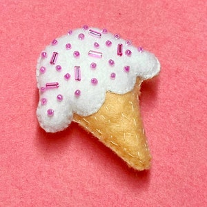 May include: A white felt ice cream cone with pink sprinkles. The cone is on a pink felt background.