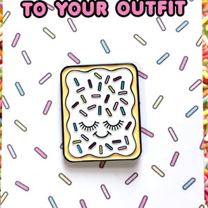 May include: A white enamel pin with a yellow outline, depicting a smiling, closed-eye Pop-Tart with colourful sprinkles. The pin is on a white card with the text "ADD A LITTLE SWEETNESS TO YOUR OUTFIT" and the website "CLAIREYLOUCREATIONS.COM".