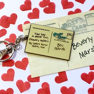 May include: A gold-coloured keychain with a postcard-shaped charm. The charm has a handwritten message that reads "Your hair is winter fire, January embers. My heart burns there, too." The postcard is addressed to Bev Marsh and Beverly Marsh.