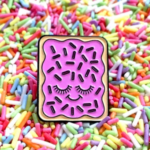 May include: A pink enamel pin shaped like a square pastry with a smiling face and sprinkles. The pin is surrounded by colourful sprinkles.