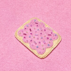 May include: A pink felt patch shaped like a square with rounded corners, decorated with pink, purple, and white beads. The patch is a felt craft of a popular breakfast pastry.