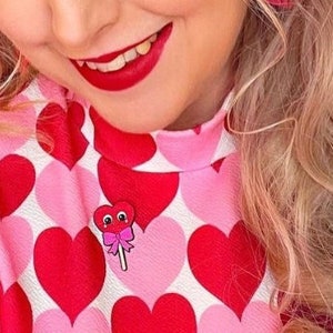 May include: A woman with pink hair wearing a pink and red heart patterned dress with a red heart lollipop pin.