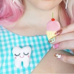 May include: A woman wearing a headband with pastel-coloured cupcake decorations, blue and pink eyeshadow, and red lipstick. She is holding a small pink cupcake with a cherry on top. She is also wearing a blue and white gingham dress with a white tooth pin.