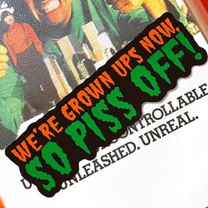 Peut inclure: Un autocollant avec le texte "WE'RE GROWN UPS NOW. SO PISS OFF!" en orange et vert sur fond noir. L'autocollant est sur un boîtier de DVD avec un homme faisant un signe de pouce. Le fond est orange.