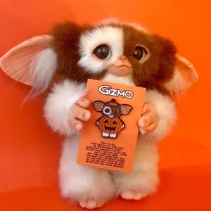 May include: A plush Gizmo figure with brown and white fur and large ears holding an orange backing card. The card features the text GIZMO and an illustration of the character inside a pumpkin, followed by the rules for caring for a Mogwai.