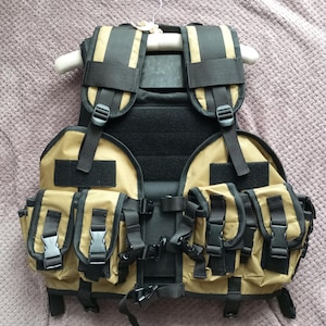 May include: A tan tactical vest with multiple black pouches and straps. The vest has a black padded back panel and is hanging on a wooden hanger.