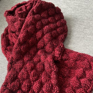 Könnte beinhalten: Ein roter Strick Schal mit einem strukturierten Muster. Der Schal ist in der Hälfte gefaltet und liegt auf einer grauen Oberfläche.