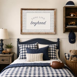 May include: A framed wall art print featuring the text "LONG LIVE boyhood" in dark blue lettering, centered on a cream background with a navy and white plaid border. The print is displayed in a rustic wooden frame above a navy blue bed frame.