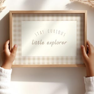 May include: A framed print with the text "Stay Curious little explorer." The print has a light beige gingham pattern and a cream-colored center. The frame is made of light-colored wood. The print is held by a person wearing a white sweater.