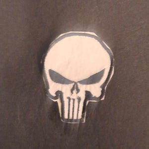 May include: A white skull patch with a black outline. The skull has a menacing expression with a prominent jawline and a large, black, stylized skull symbol on the forehead.