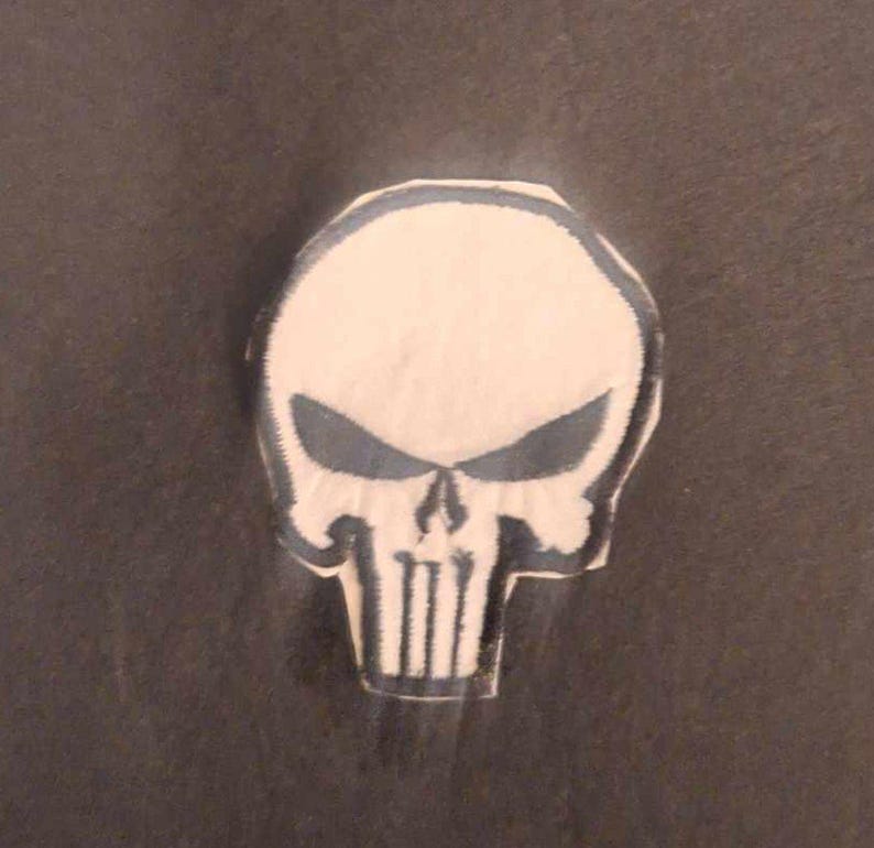 May include: A white skull patch with a black outline. The skull has a menacing expression with a prominent jawline and a large, black, stylized skull symbol on the forehead.