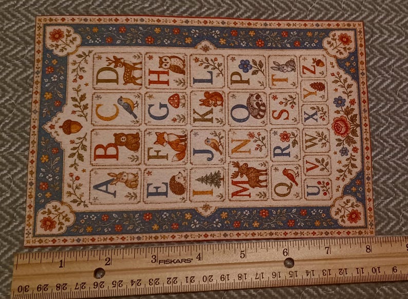 May include: A rectangular alphabet card featuring colorful letters A through Z paired with woodland animal and nature illustrations, bordered by a floral pattern. A wooden ruler placed below shows the card is approximately 8 inches wide.