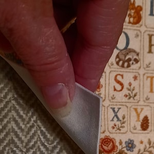 May include: A fabric sheet featuring a grid of alphabet letters, each paired with small illustrations of animals, plants, and flowers. The material has a smooth, light-colored backing visible where the corner is folded over, revealing a textured surface.