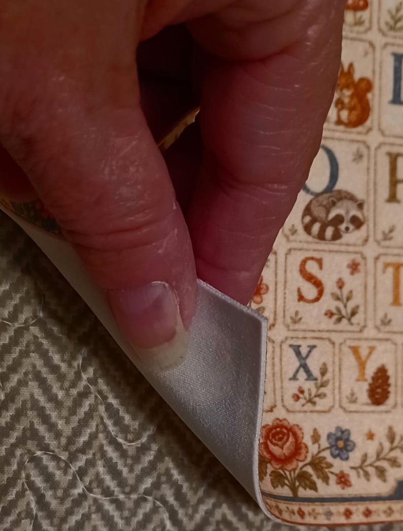 May include: A fabric sheet featuring a grid of alphabet letters, each paired with small illustrations of animals, plants, and flowers. The material has a smooth, light-colored backing visible where the corner is folded over, revealing a textured surface.