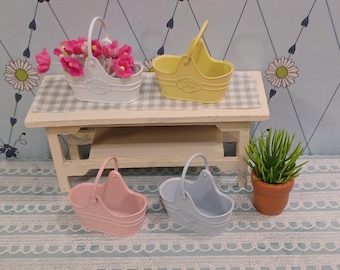 May include: A collection of miniature metal baskets in pastel colors: white, yellow, pink, and blue. One basket holds pink flowers. The baskets have a handle and decorative detailing. A small potted plant sits nearby.