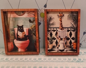 May include: Two framed art prints. One features a black cat reading on a toilet in a pink bathroom. The other shows a giraffe wearing glasses, reading a newspaper on a toilet in a black and white tiled bathroom.