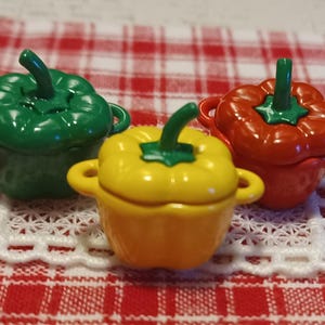 May include: Three miniature lidded pots shaped like bell peppers in green, yellow, and red. Each pot features a small stem-shaped handle on the lid and two side handles, with a glossy finish and a smooth, rounded body design.