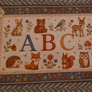 May include: A rectangular woven mat featuring the letters ABC in the center, surrounded by illustrations of a rabbit, bear, bird, deer, hedgehog, fox, squirrel, and owl, with floral accents and a dark blue border with orange and white flowers.