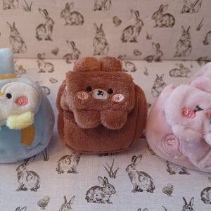 May include: Three plush animal-shaped coin purses. The blue purse is a penguin, the brown one is a bear, and the pink one is a bunny. Each has embroidered details and a small handle or strap. The background is a fabric with a rabbit pattern.
