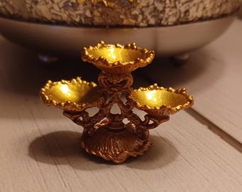 May include: A gold-tone metal decorative stand with three tiers. Each tier has a round, scalloped edge. The stand has an ornate design with intricate details.