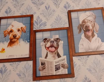 May include: Three rectangular framed wall art pieces featuring dogs in bathroom settings. One shows a dog in a bubble bath with a rubber duck, another a dog on a toilet reading a newspaper titled Daily Doo, and the third a dog in a white bathrobe.