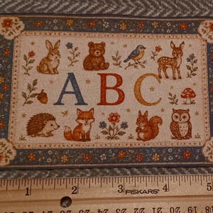 May include: A rectangular card featuring a woodland animal illustration with the letters ABC in the center. The design includes a rabbit, bear, bird, deer, hedgehog, fox, squirrel, and owl, bordered by a floral pattern. A wooden ruler shows a width of 6 inches.