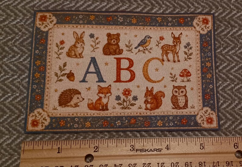 May include: A rectangular card featuring a woodland animal illustration with the letters ABC in the center. The design includes a rabbit, bear, bird, deer, hedgehog, fox, squirrel, and owl, bordered by a floral pattern. A wooden ruler shows a width of 6 inches.