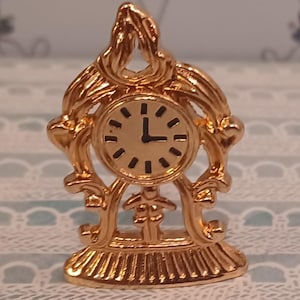 May include: A small, gold-colored decorative clock. The clock face is round with black numerals and hands, indicating the time is approximately 2:00. The clock has an ornate, openwork design with a base and a decorative top.