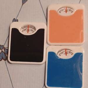 May include: Three miniature square bathroom scales in black, peach, and blue. Each scale features a white frame, a circular analog dial with a red needle, and markings ranging from 0 to 120, with the text 10kg capacity printed on the dial face.