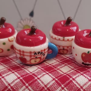May include: Four miniature apple-shaped mugs with red, glossy tops and brown stems. The mugs are white with apple designs and blue handles. One mug has the word "Apple" on it.