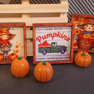 May include: Three square wooden signs with autumn themes featuring a scarecrow, a green truck with pumpkins, and text reading Fall Y'all and Farm Fresh Pumpkins, accompanied by four small orange pumpkin figurines with green stems.