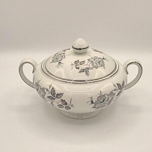 May include: A white porcelain sugar bowl with a lid, featuring a gray floral pattern of roses and leaves. The bowl has two curved handles and a silver-toned trim along the rim, the lid edge, the handle edges, and the lid finial knob.